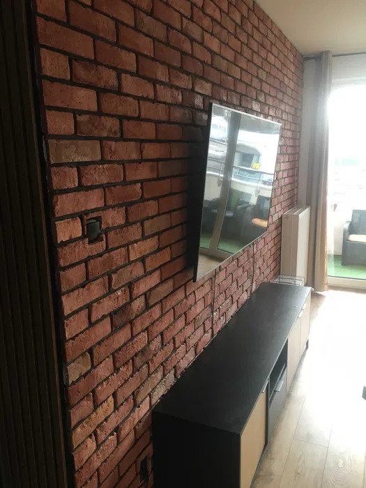 Wykończona ściana z cegły dekoracyjnej w salonie z telewizorem. Widoczna komoda RTV i fragment okna balkonowego z zasłoną.