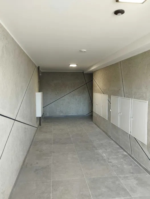 Korytarz w budynku z szarymi płytkami podłogowymi i dekoracyjnymi ścianami z geometrycznym wzorem. Białe szafki na ścianie po prawej stronie.