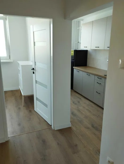 Jasne wnętrze po remoncie: panele podłogowe, białe ściany, drzwi i meble. Widok na kuchnię z szarymi szafkami i drewnianym blatem.