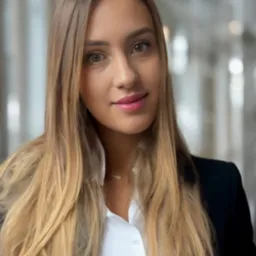 Portret uśmiechniętej kobiety w białej koszuli i czarnej marynarce na rozmytym tle biurowym.