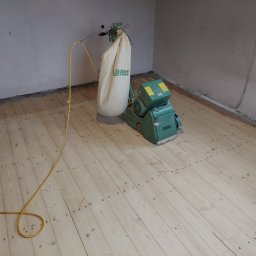 Michał Dobija - Maszyna do renowacji drewnianych podłóg firmy z Łodygowic, z workiem na pył marki Lägler, stoi na jasnej, wyszlifowanej podłodze w pokoju z surowymi ścianami.