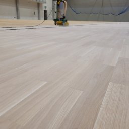 Michał Dobija - Maszyna do cyklinowania w trakcie pracy na jasnej drewnianej podłodze w hali sportowej.