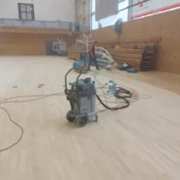 Cyklinowanie parkietu w hali sportowej, widoczny sprzęt Bona i pracownik w tle.