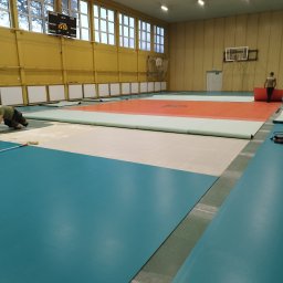 Michał Dobija - Montaż sportowej podłogi w hali: pracownik rozprowadza klej wałkiem, widoczne rozłożone fragmenty podłogi w kolorach niebieskim, beżowym i pomarańczowym, w tle tablica wyników i kosz do koszykówki.