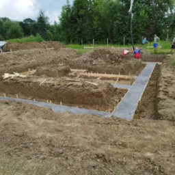 Wykopane fundamenty z drewnianym szalunkiem, częściowo wypełnione betonem, widoczna pompa do betonu i pracownicy w tle.
