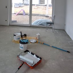 Michał Dobija - Wnętrze w trakcie malowania: wiaderka z farbą, kuweta malarska z wałkiem, teleskopowy uchwyt do wałka, oraz krzesło w tle, widok przez okno na plac budowy z dziecięcą zjeżdżalnią.