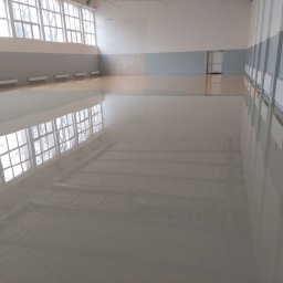 Michał Dobija - Jasnoszara, błyszcząca posadzka żywiczna w hali sportowej z dużymi oknami i grzejnikami przy ścianach.