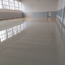 Jasnoszara, błyszcząca posadzka żywiczna w hali sportowej z dużymi oknami i grzejnikami przy ścianach.