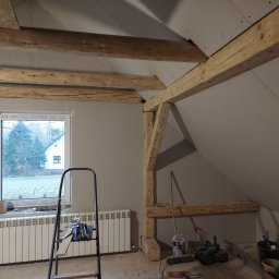 Michał Dobija - Poddasze w trakcie remontu: surowe drewniane belki stropowe, ściany wyłożone płytami gipsowo-kartonowymi, grzejnik pod oknem, drabina i narzędzia budowlane na podłodze.