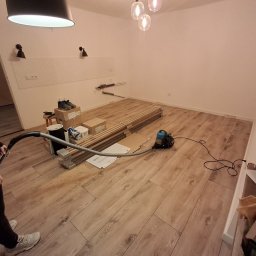 Michał Dobija - Kobieta odkurza nowo ułożoną podłogę z paneli, na podłodze leżą opakowania po panelach i narzędzia.