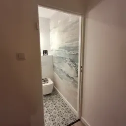 Wąska toaleta z tapetą w morskim stylu, białą ceramiką, wzorzystą podłogą i parą butów na desce sedesowej, widziana z korytarza z drewnianą podłogą.