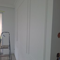 Michał Dobija - Świeżo pomalowana ściana z dekoracyjnymi panelami i listwami, widoczna drabina malarska i niedokończone gniazdka elektryczne.