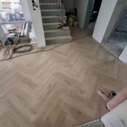 Jasna podłoga ułożona w jodełkę, widok z góry. W tle schody, narzędzia i materiały budowlane. Częściowo widoczna rolka wykładziny.