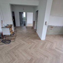 Michał Dobija - Jasne panele ułożone we wzór jodełki francuskiej w przestronnym, odświeżanym wnętrzu. Widoczne przygotowania do dalszych prac wykończeniowych, narzędzia i materiały.