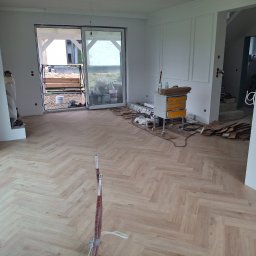 Michał Dobija - Ułożona parkietowa podłoga w jodełkę w jasnym pomieszczeniu z białymi ścianami i otwartym przejściem. Widoczne narzędzia i materiały budowlane.