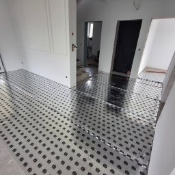 Michał Dobija - Rozłożona folia grzewcza na podłodze w jasnym wnętrzu, przygotowana do dalszych prac wykończeniowych. Widoczne białe ściany i drzwi w trakcie remontu.