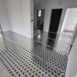 Rozłożona folia grzewcza na podłodze w jasnym wnętrzu, przygotowana do dalszych prac wykończeniowych. Widoczne białe ściany i drzwi w trakcie remontu.