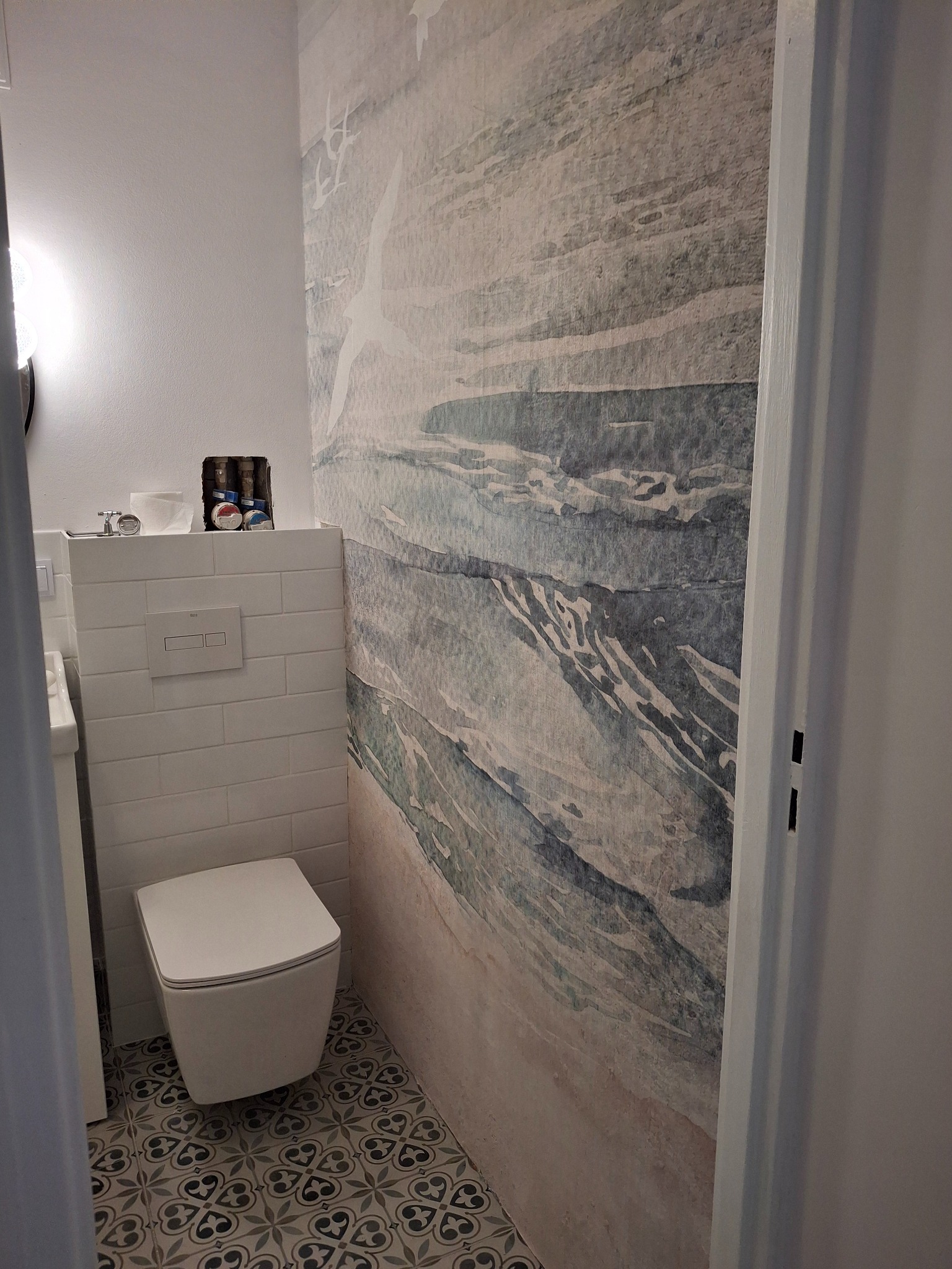 Wnętrze łazienki z toaletą podwieszaną, płytkami imitującymi cegłę, podłogą w stylu retro i tapetą z motywem morskim.