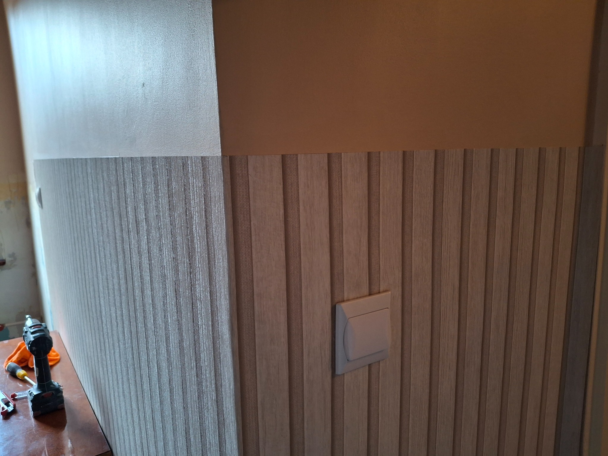 Montaż paneli ściennych z drewnopodobnym wzorem w pionowe pasy. Widoczny włącznik światła na ścianie oraz narzędzia pracy: wkrętarka i śrubokręty na stole.