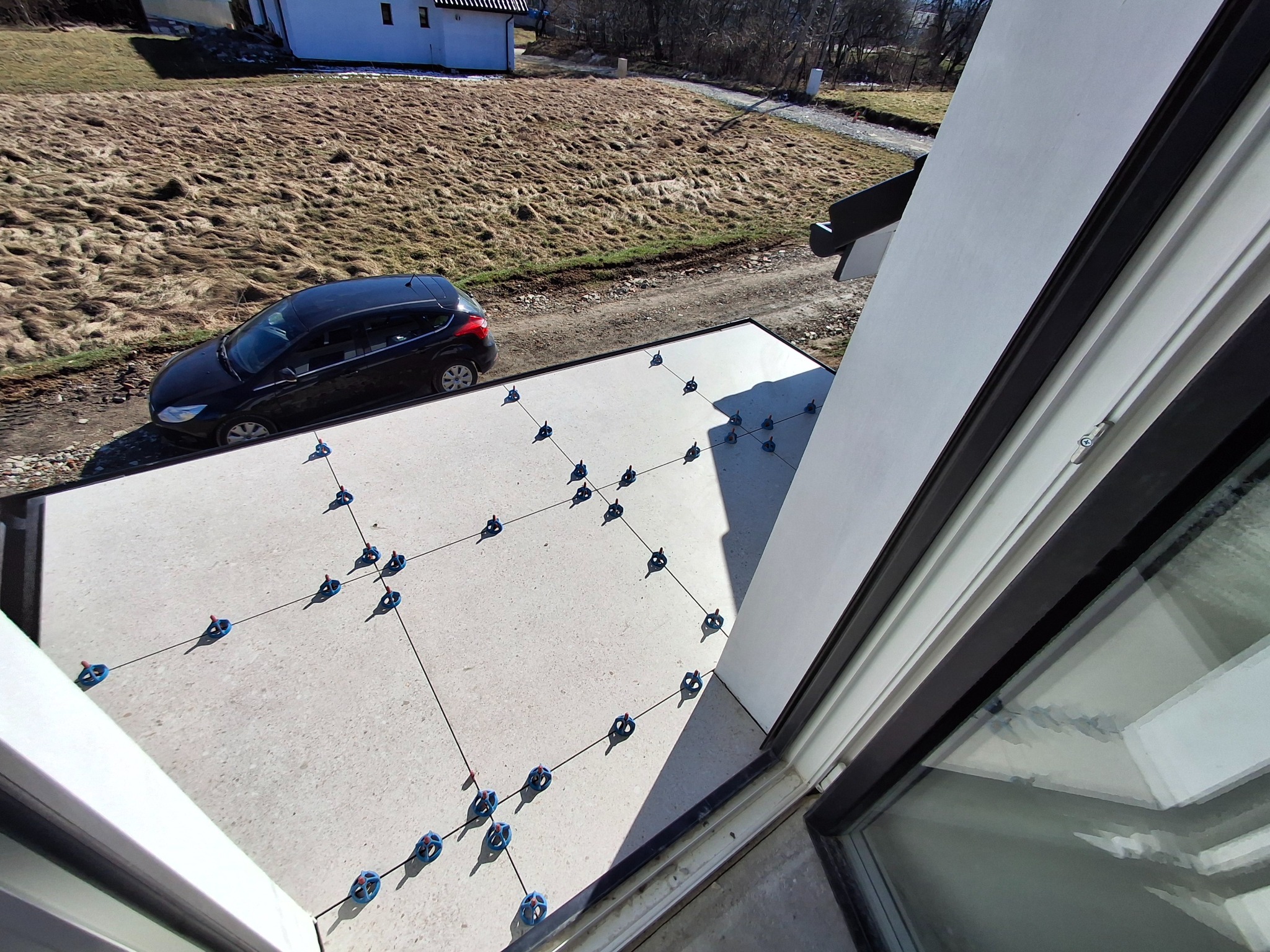 Montaż płyt tarasowych na balkonie z użyciem systemu poziomowania, widok z okna na samochód i dom w tle.