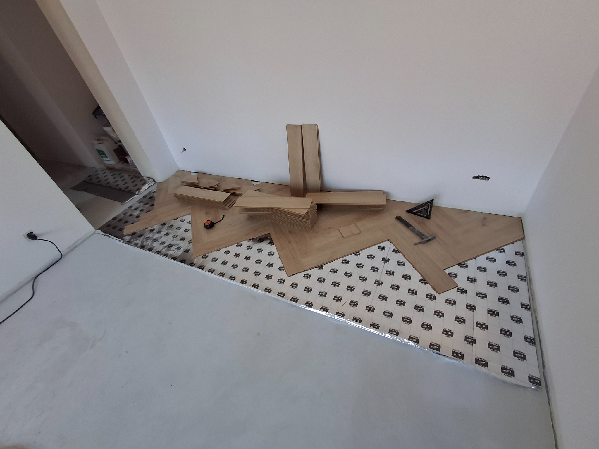 Układanie paneli podłogowych w jodełkę na podkładzie izolacyjnym, widoczne narzędzia: miarka, kątownik i młotek.