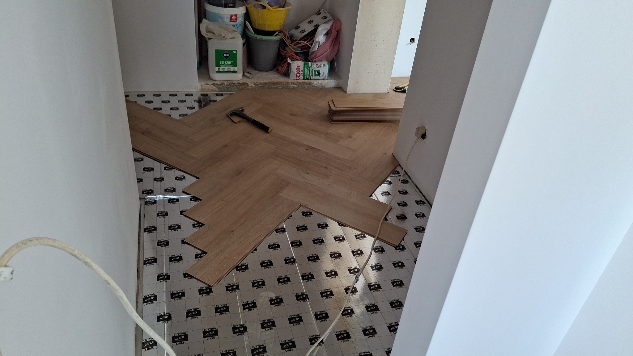 Podłoga w trakcie wykańczania panelami ułożonymi we wzór jodełki, widoczny młotek, pozostałe panele i podkład pod panele z nadrukiem, w tle puszki z farbami i zaprawa.