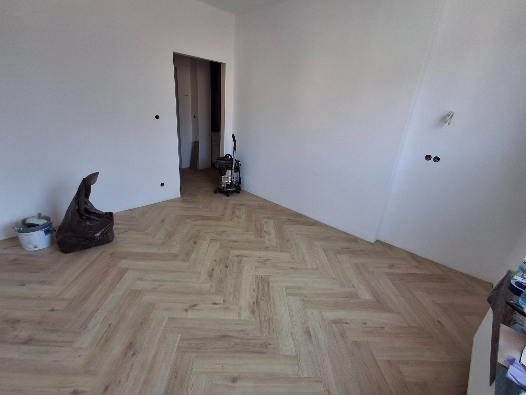 Jasne panele ułożone w jodełkę francuską w pustym, świeżo wykończonym pokoju z białymi ścianami. Widoczny odkurzacz przemysłowy, wiadro i worek na śmieci.