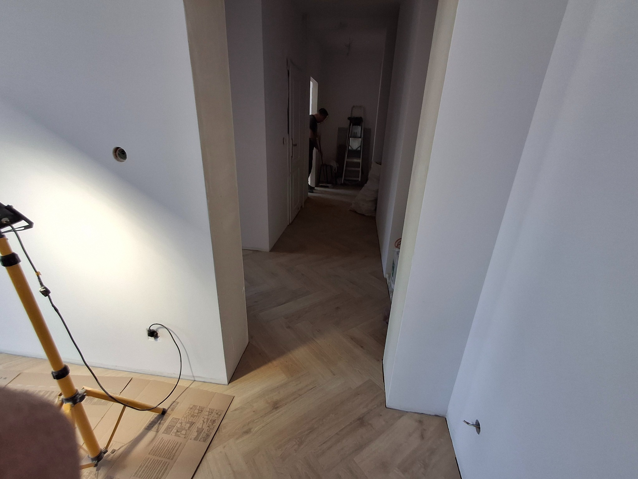 Korytarz w trakcie wykańczania z ułożoną drewnianą podłogą w jodełkę francuską, widoczna lampa budowlana i instalacja elektryczna bez osprzętu, w tle osoba pracująca w oddali.