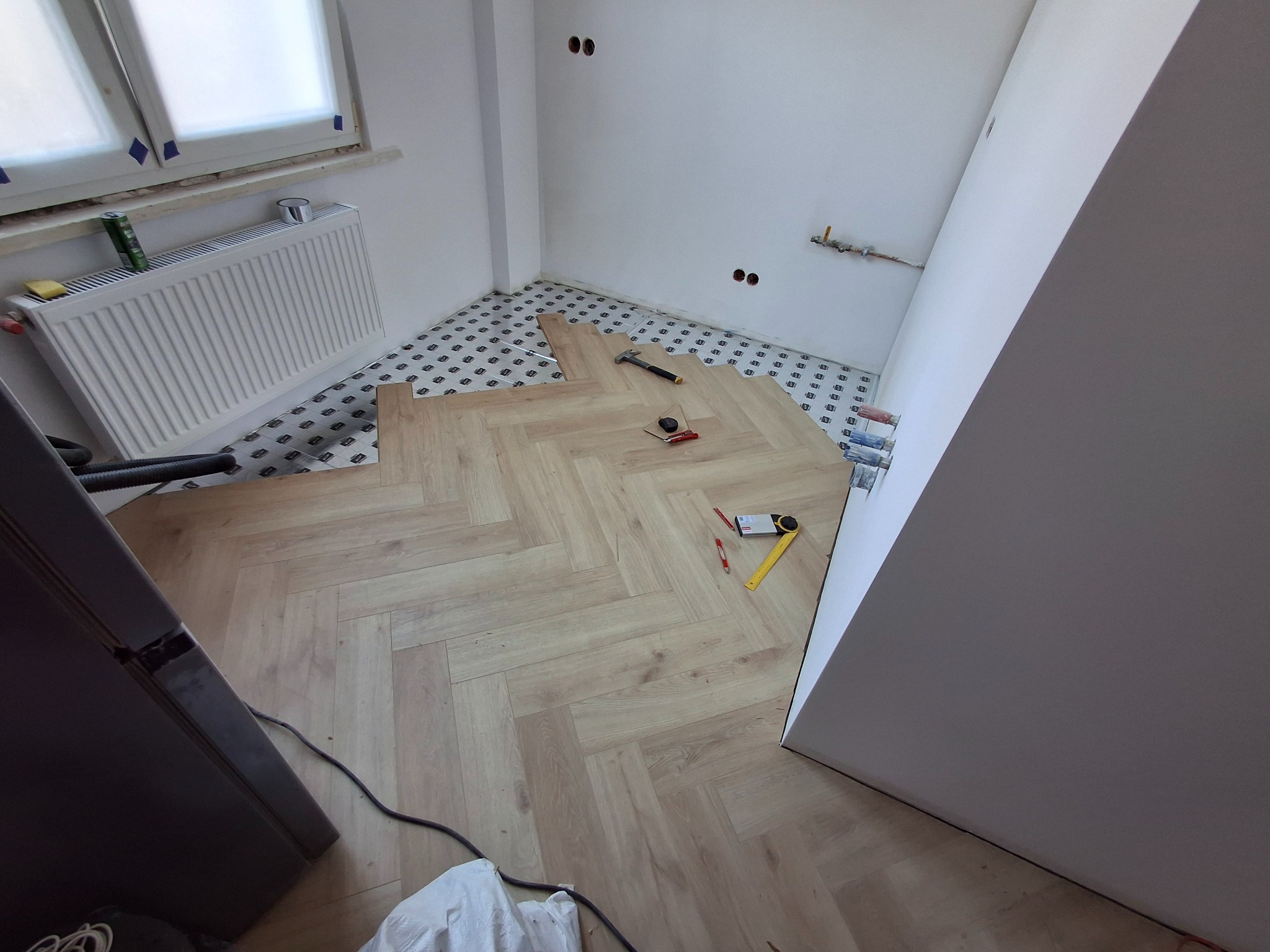 Wnętrze w trakcie prac wykończeniowych: panele ułożone we wzór jodełki na podkładzie, widoczne narzędzia pomiarowe i młotek, ściany przygotowane do malowania, grzejnik pod oknem.