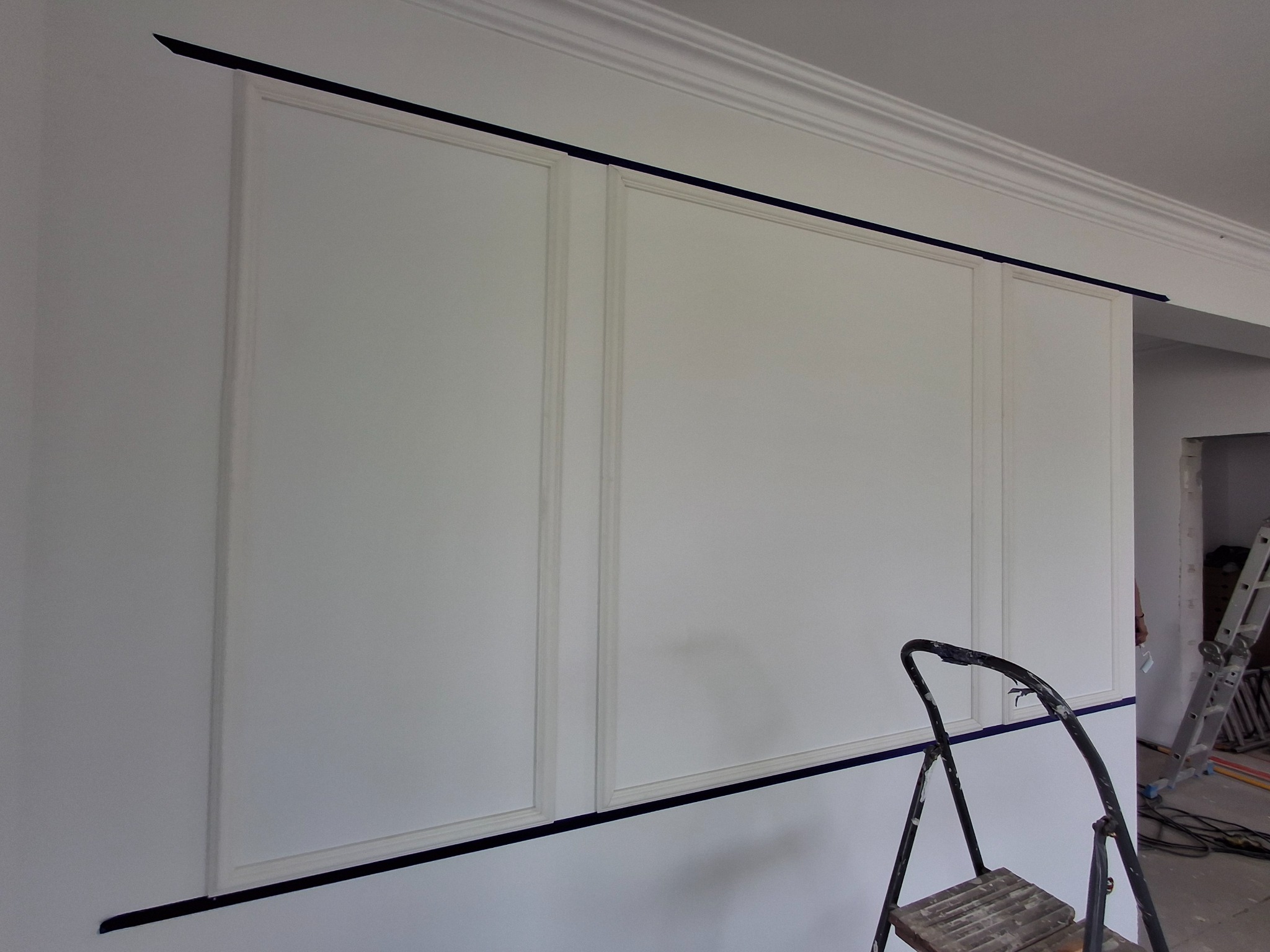 Ściana z białymi panelami dekoracyjnymi, zabezpieczona taśmą malarską, z fragmentem drabiny malarskiej w prawym dolnym rogu, w tle widoczny remont.