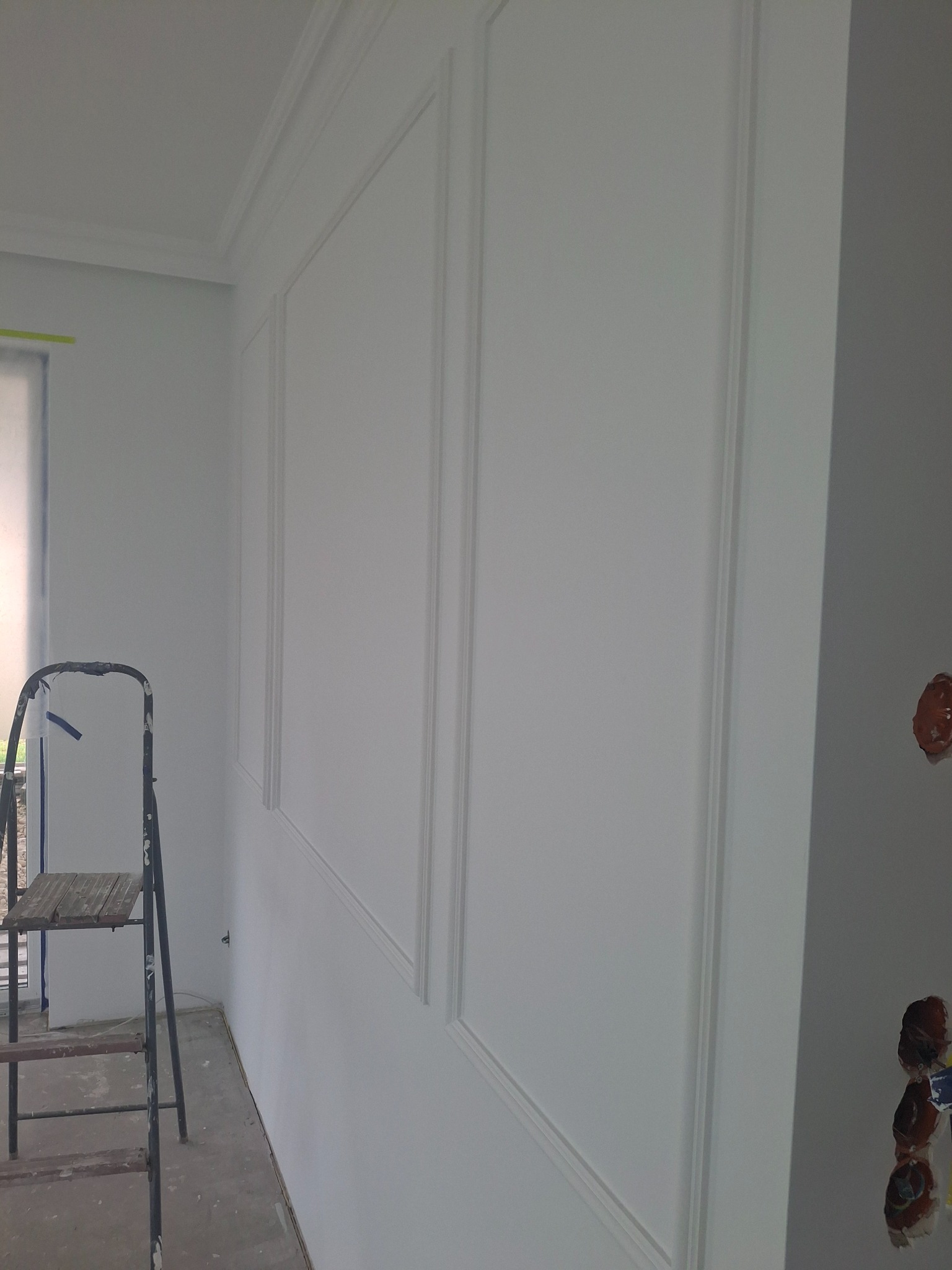 Świeżo pomalowana ściana z dekoracyjnymi panelami i listwami, widoczna drabina malarska i niedokończone gniazdka elektryczne.