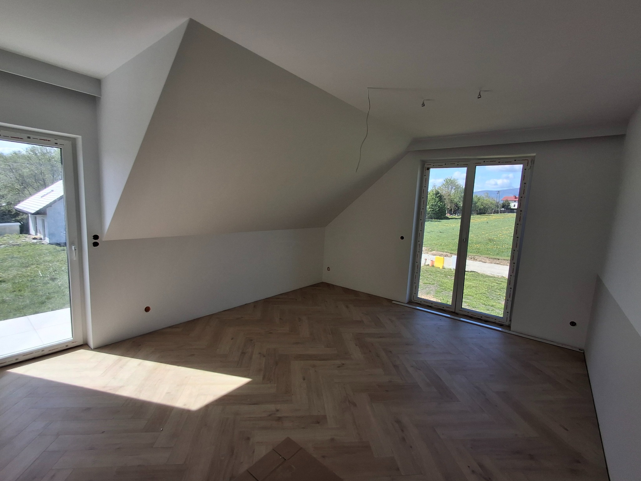 Jasne panele ułożone w jodełkę w pokoju na poddaszu z oknami. Świeżo pomalowane ściany w odcieniach bieli. Widok na zewnątrz przez okna.