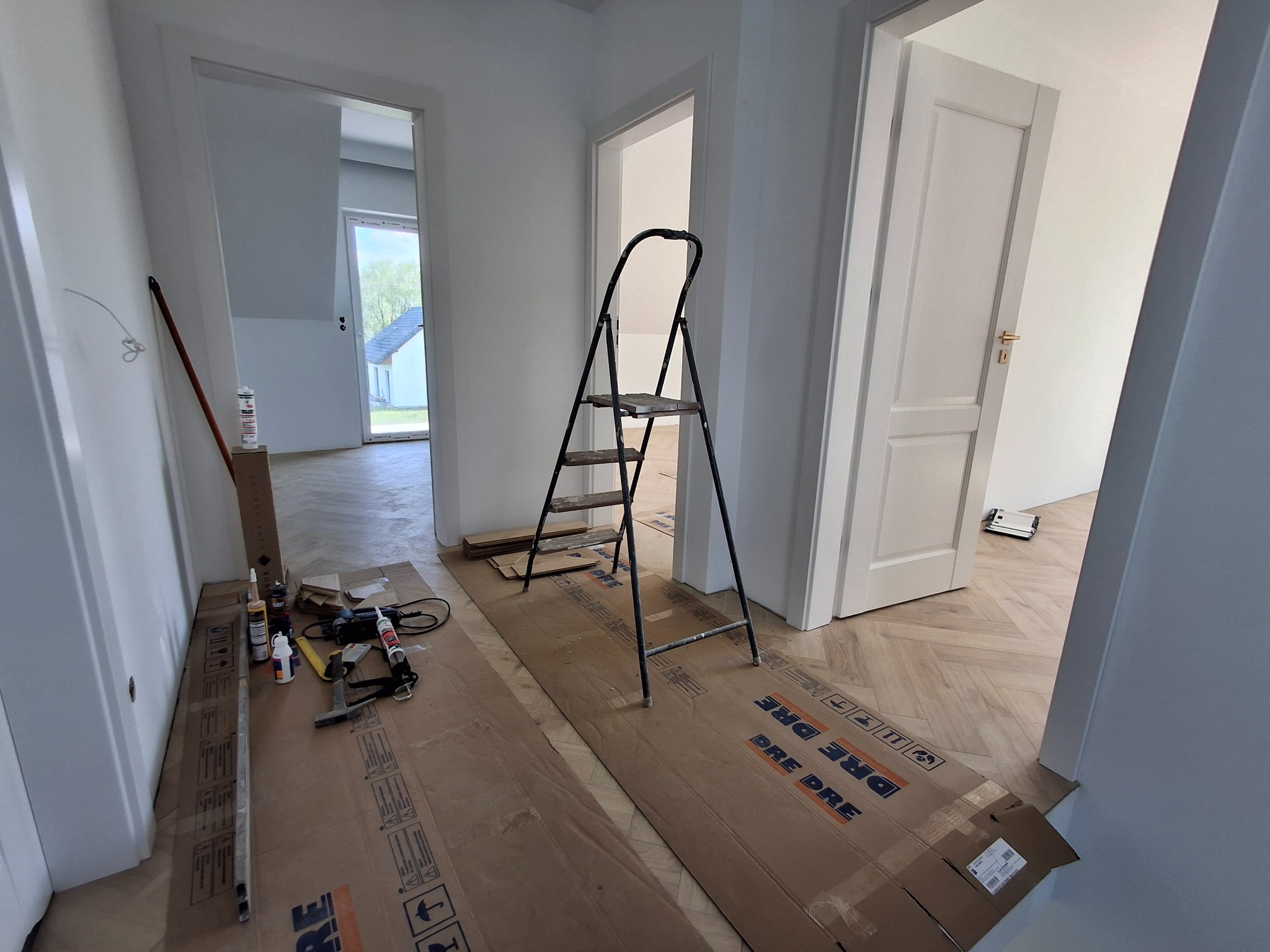 Wnętrze w trakcie prac wykończeniowych: parkiet w jodełkę zabezpieczony kartonem, drabina, narzędzia i materiały budowlane przy ścianie. Widoczne ościeżnice i drzwi.