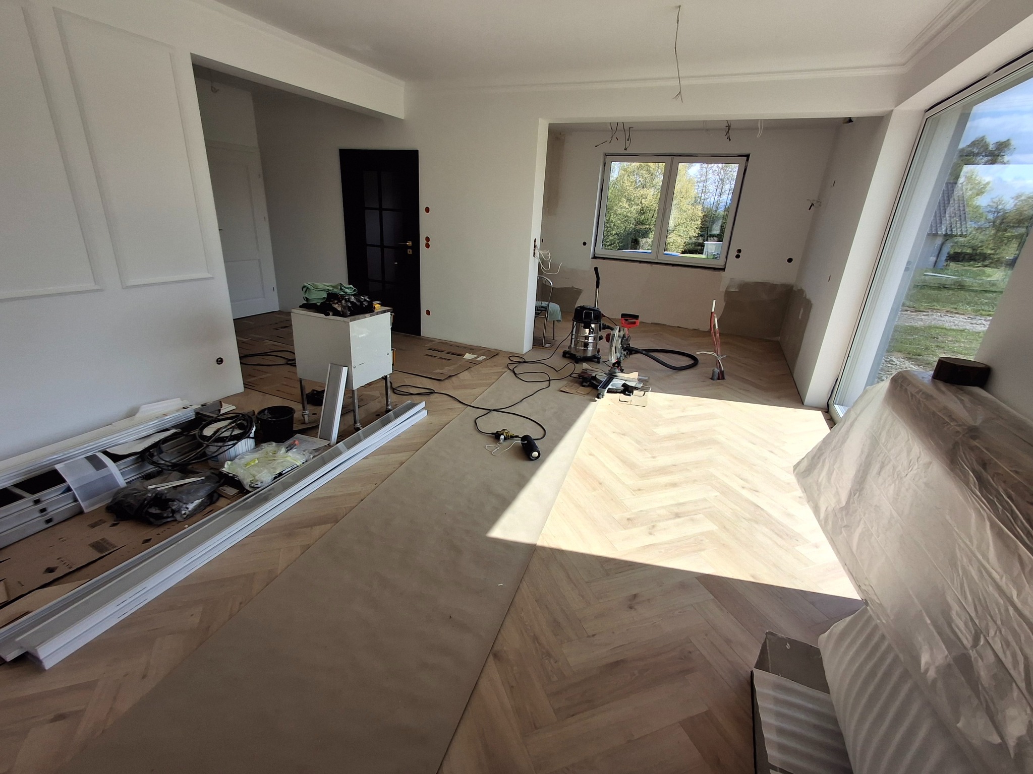 Wnętrze w trakcie remontu: parkiet w jodełkę, narzędzia, listwy przypodłogowe. Widoczne okno i drzwi w tle. Częściowo zakryte meble folią ochronną.