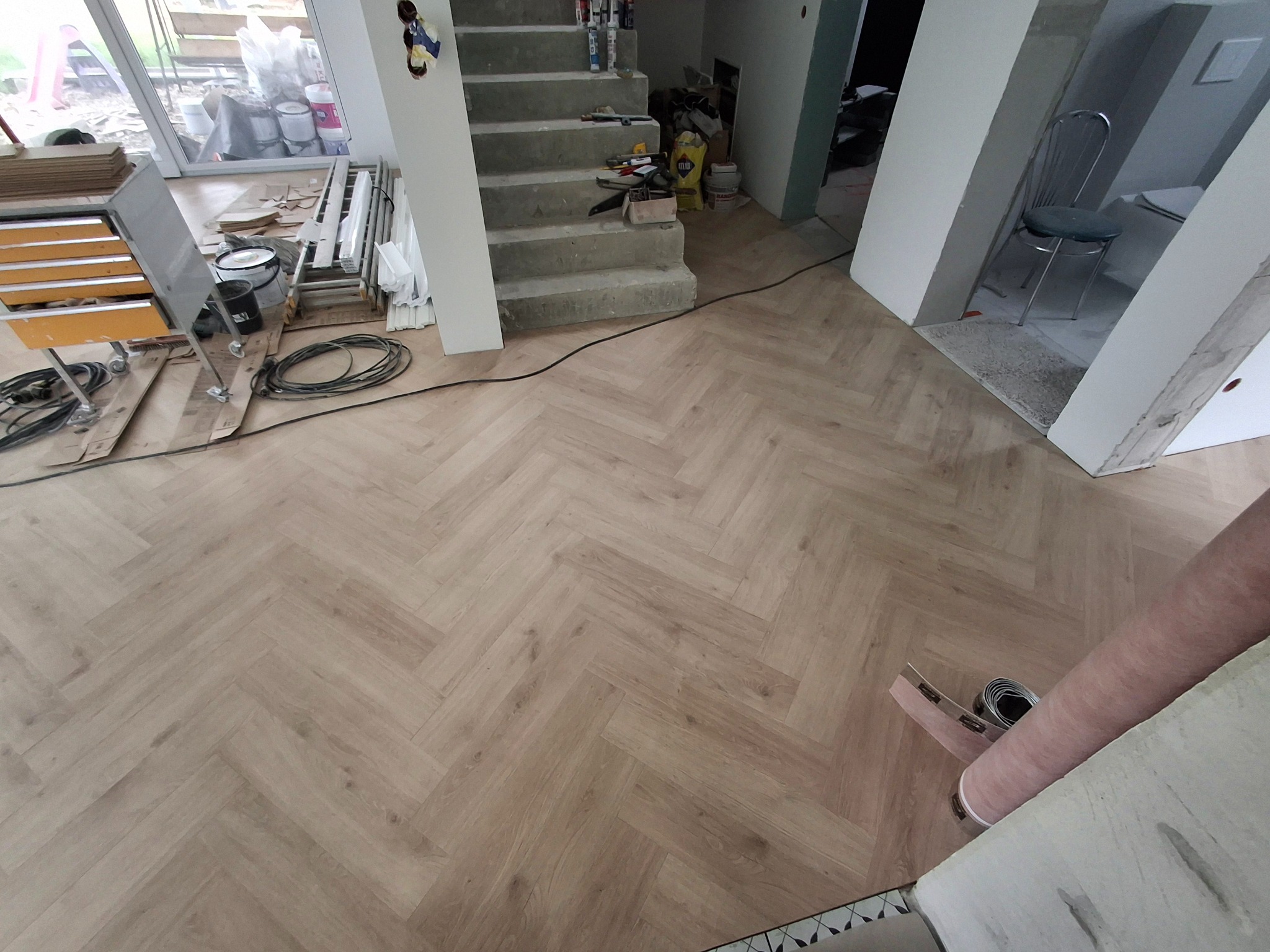 Jasna podłoga ułożona w jodełkę, widok z góry. W tle schody, narzędzia i materiały budowlane. Częściowo widoczna rolka wykładziny.