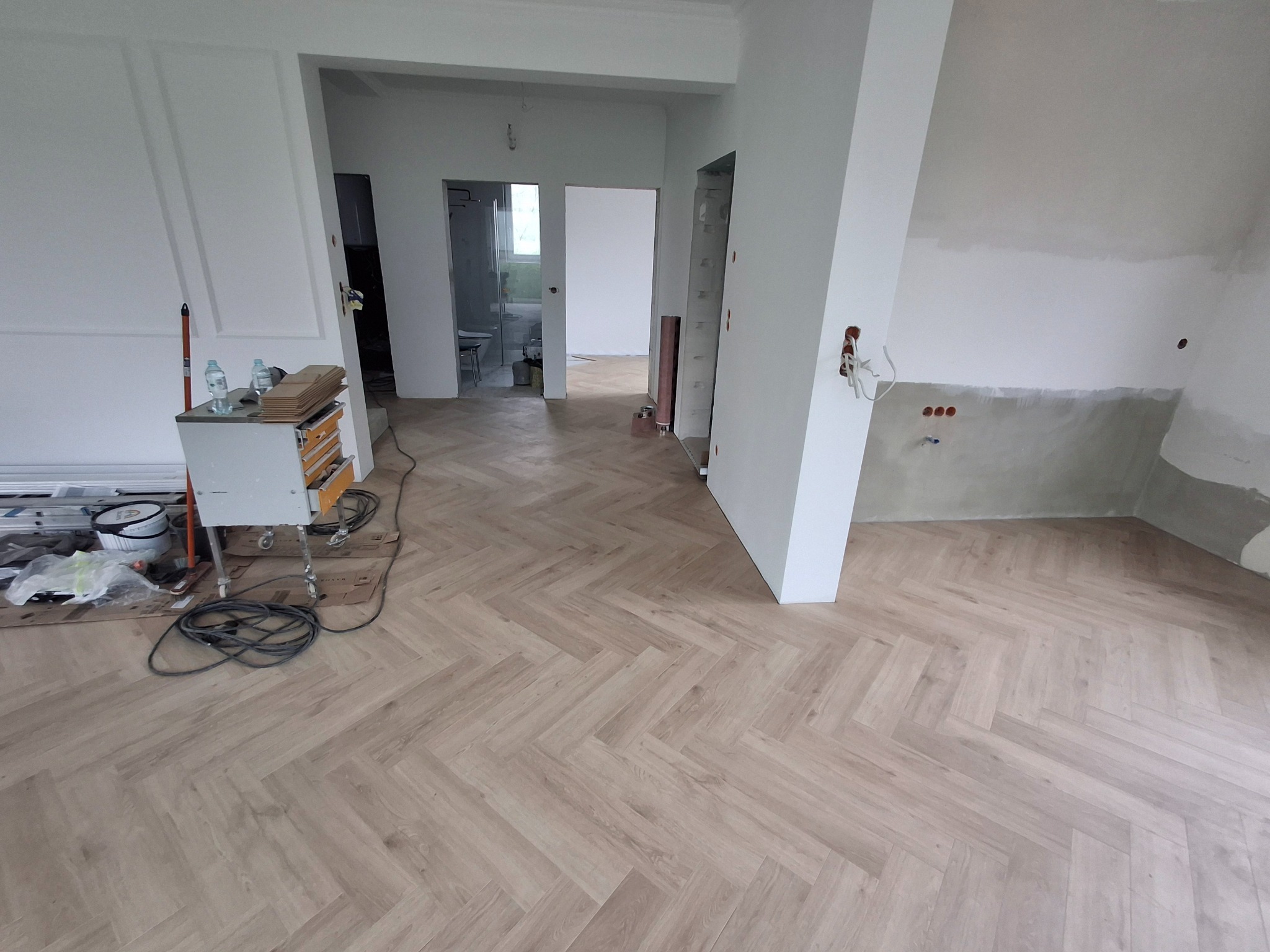 Jasne panele ułożone we wzór jodełki francuskiej w przestronnym, odświeżanym wnętrzu. Widoczne przygotowania do dalszych prac wykończeniowych, narzędzia i materiały.