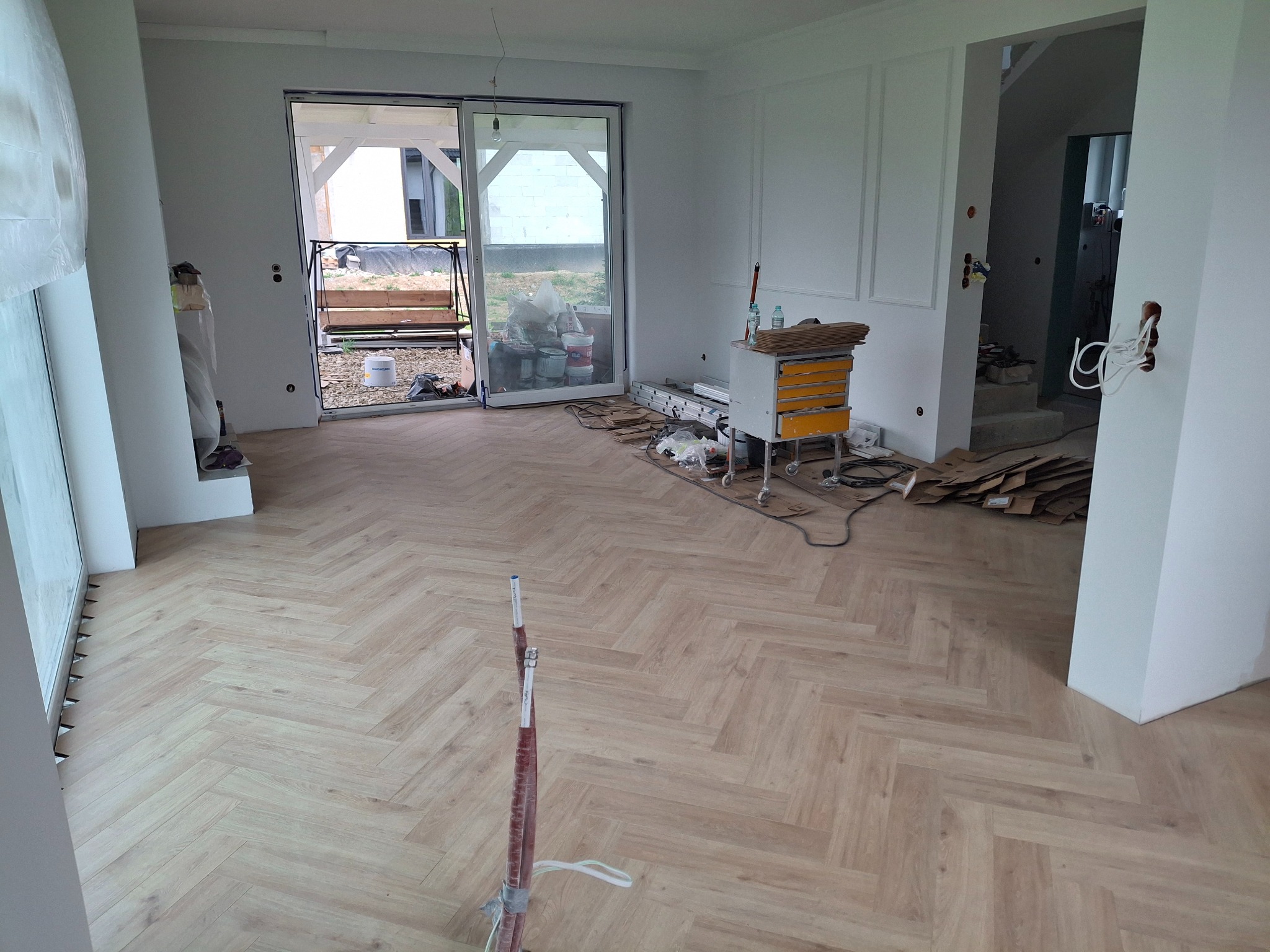 Ułożona parkietowa podłoga w jodełkę w jasnym pomieszczeniu z białymi ścianami i otwartym przejściem. Widoczne narzędzia i materiały budowlane.