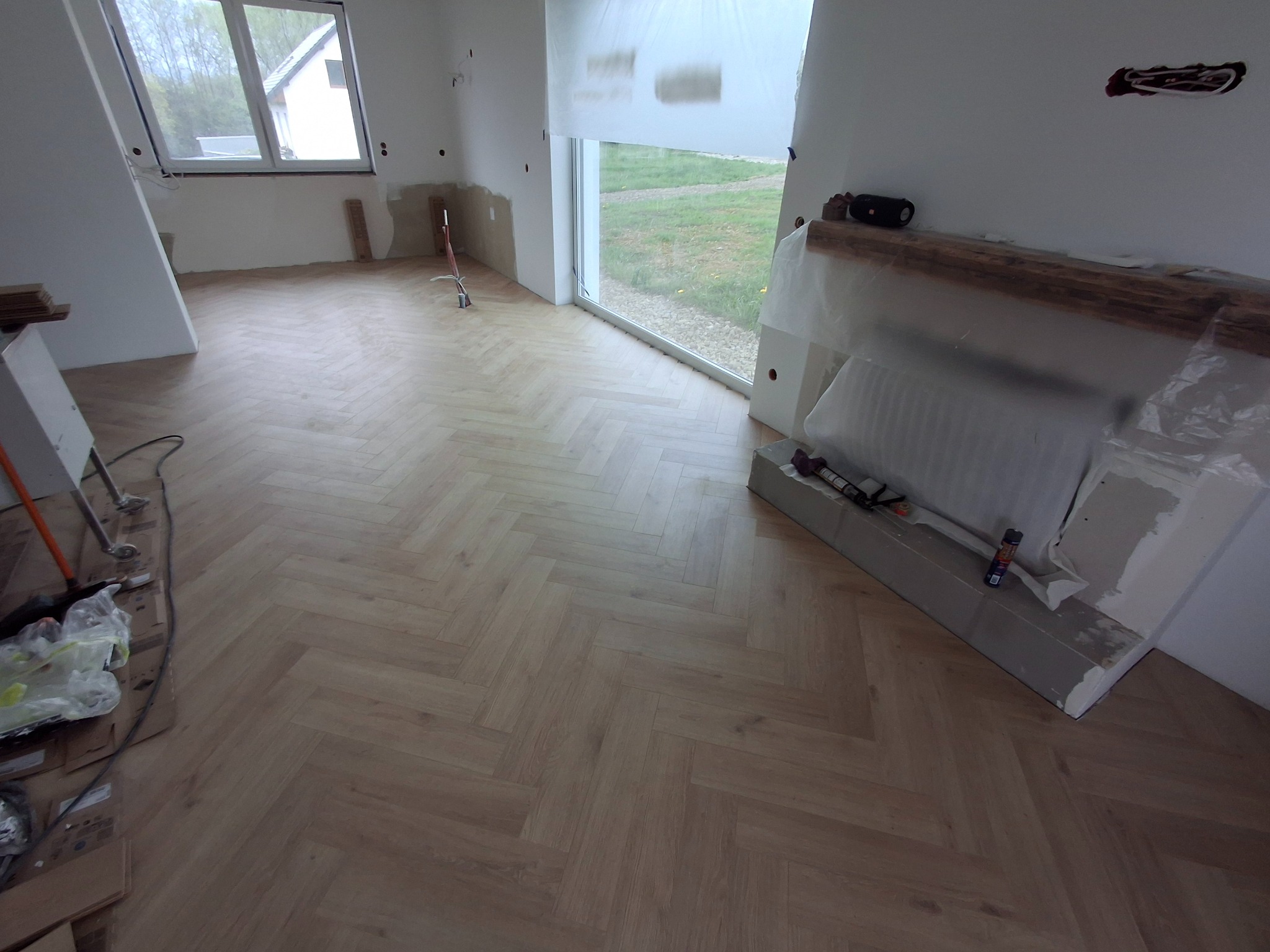 Jasna podłoga z paneli ułożonych w jodełkę w nowym wnętrzu. Widoczne okno i przygotowane ściany do wykończenia. Część materiałów i narzędzi w rogu pomieszczenia.