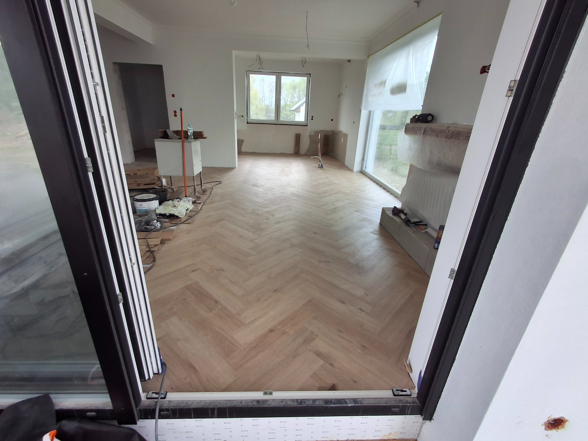 Jasne panele ułożone w jodełkę w przestronnym, jasnym pokoju. Widok z perspektywy otwartych drzwi balkonowych. W tle okno i sprzęty zakryte folią.