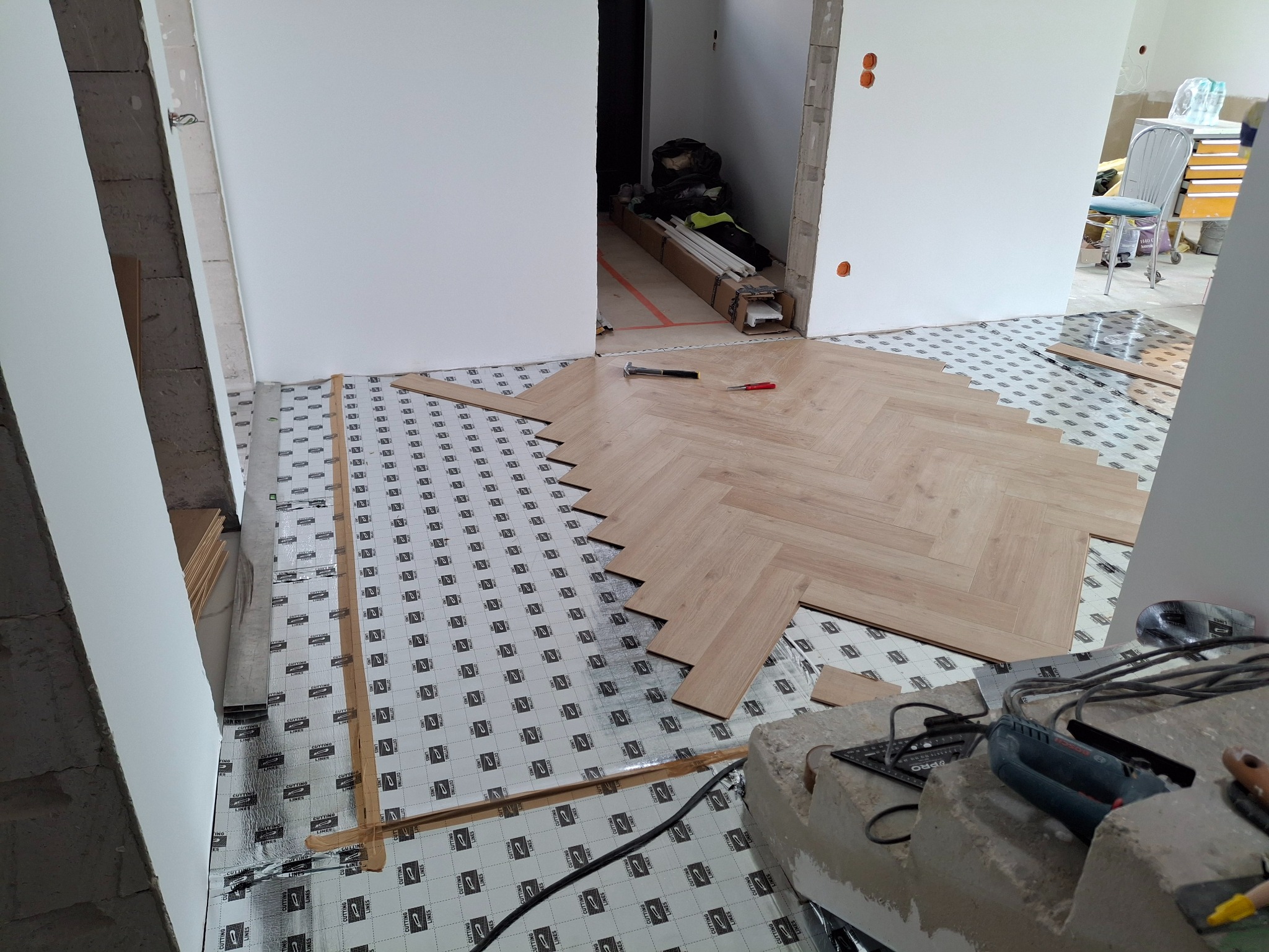 Układanie paneli podłogowych w jodełkę, widoczne narzędzia i podkład. Częściowo ułożona podłoga z jasnego drewna, w tle widoczne kolejne pomieszczenie w trakcie prac wykończeniowych.