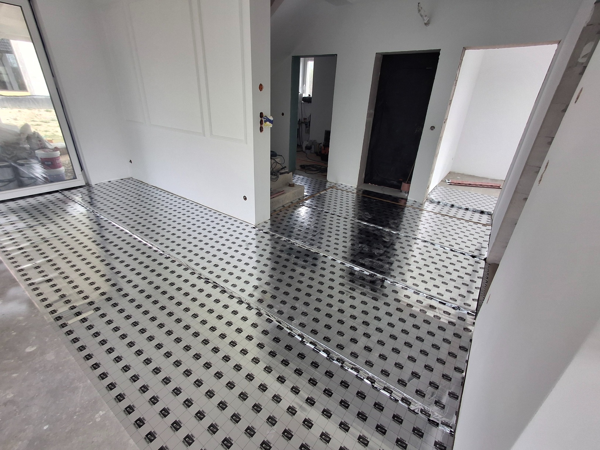 Rozłożona folia grzewcza na podłodze w jasnym wnętrzu, przygotowana do dalszych prac wykończeniowych. Widoczne białe ściany i drzwi w trakcie remontu.