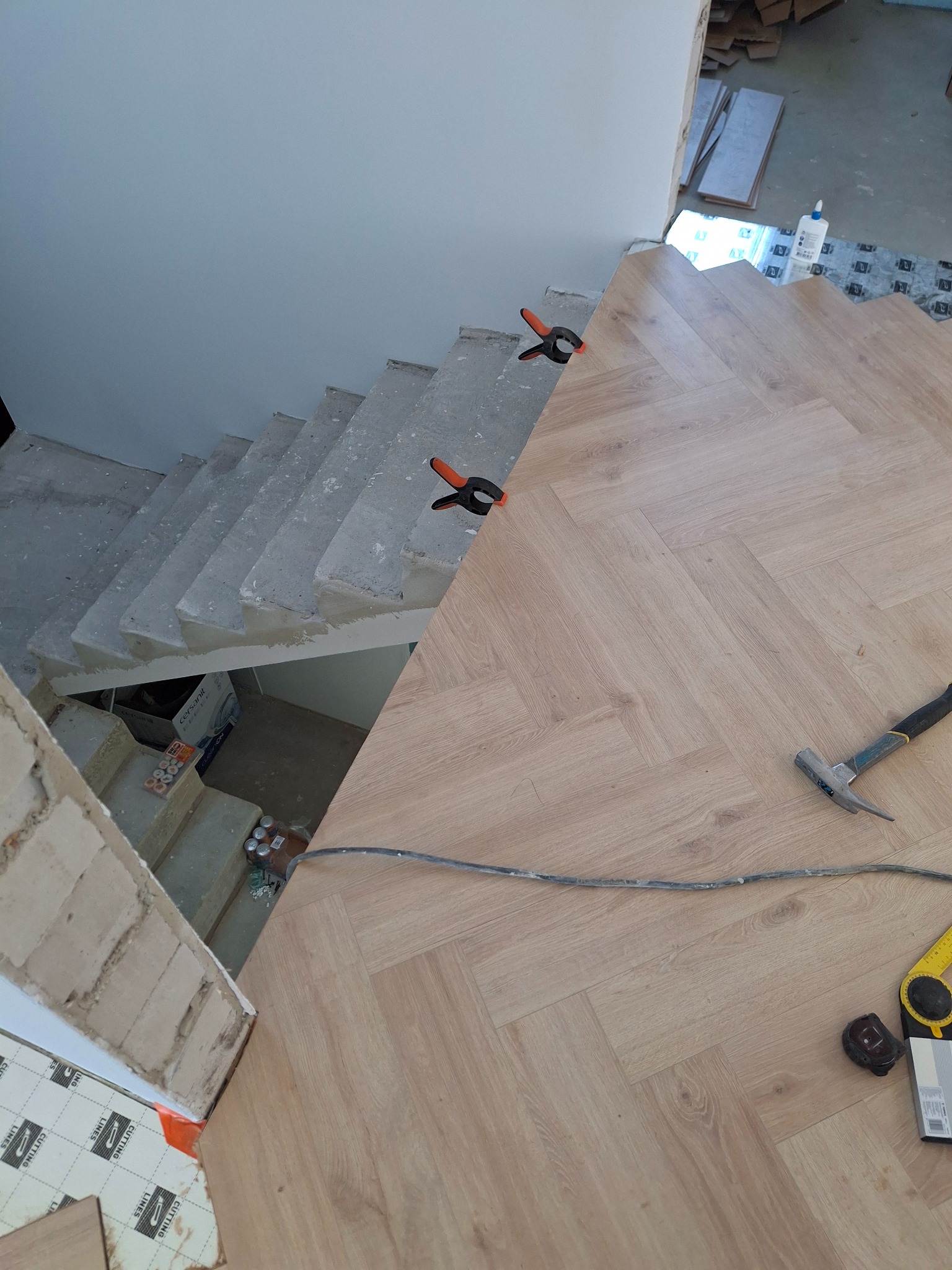 Układanie paneli w jodełkę na podłodze przy schodach. Widoczne narzędzia: młotek, miarka, kable. Fragment schodów w stanie surowym. Użyte klamry do mocowania paneli.