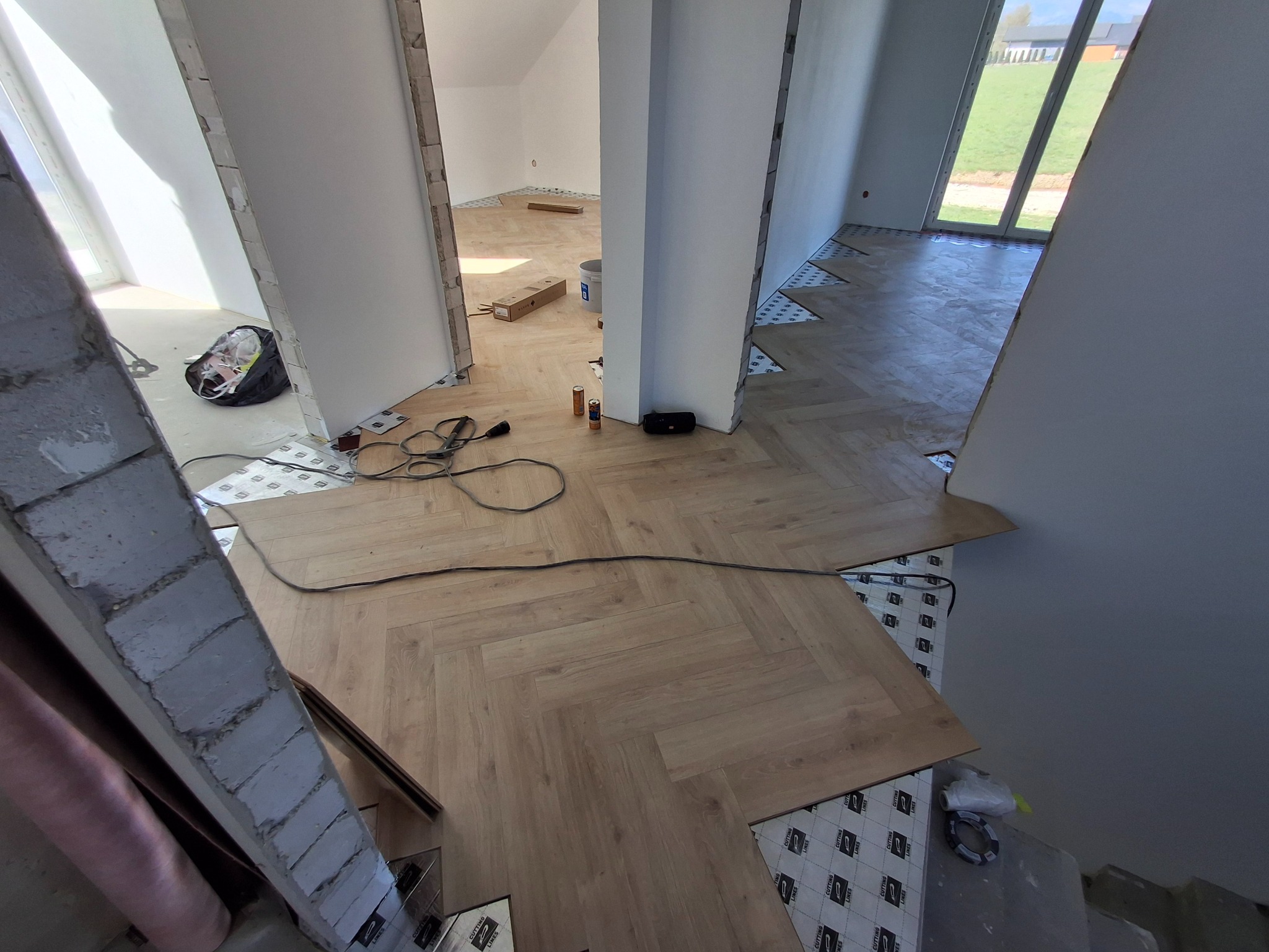 Układanie paneli w jodełkę na podkładzie izolacyjnym. Widoczny fragment podłogi z panelami, narzędzia i materiały budowlane. W tle okno i drzwi.