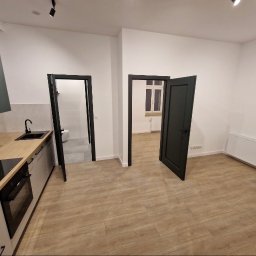 PERFECT WALLS KAMIL SEREMAK - Jasne wnętrze po remoncie: kuchnia z drewnianym blatem, widok na łazienkę i pokój przez otwarte, czarne drzwi. Minimalistyczny design, drewniana podłoga.
