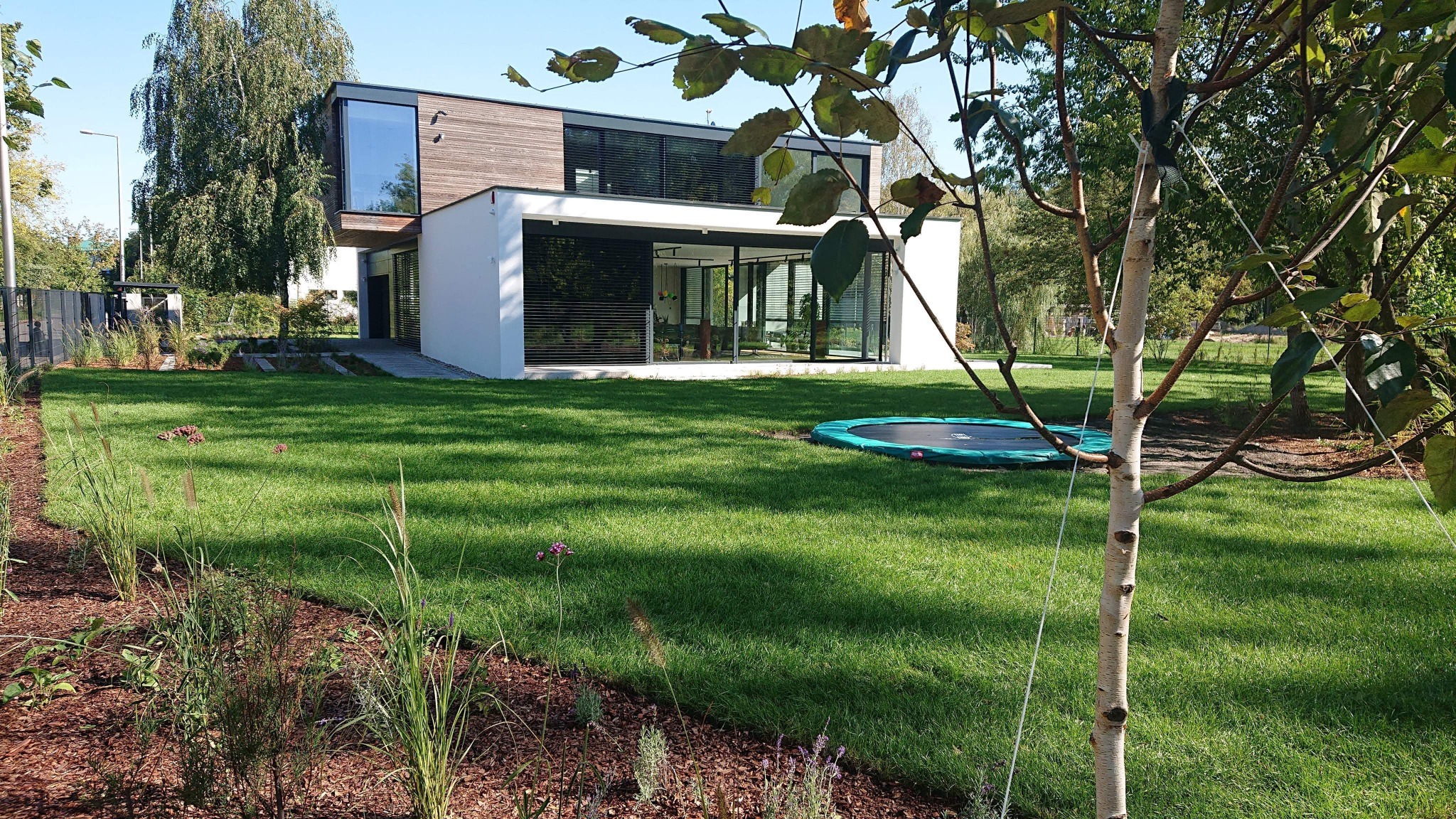 Nowoczesny dom z drewnianą elewacją i dużymi przeszkleniami, widok z ogrodu z trampoliną, Konstancin.