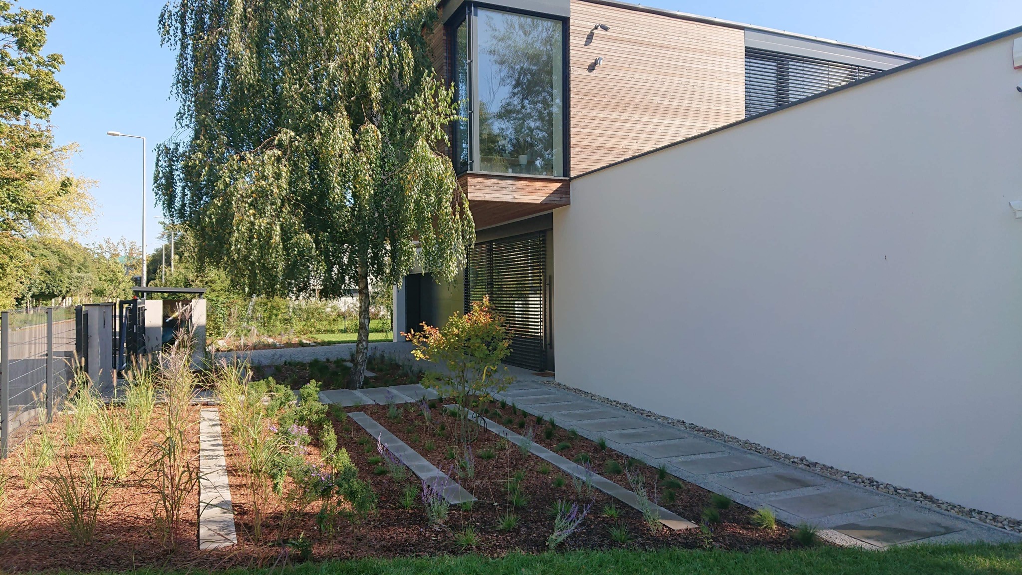 Nowoczesny dom z elewacją z jasnego drewna i białego tynku, z dużym oknem narożnym, otoczony starannie zaprojektowanym ogrodem z kamiennymi ścieżkami i ozdobnymi trawami.