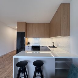 Meble na wymiar Bartosz Dopierała - Nowoczesna kuchnia z drewnianymi szafkami, marmurowym blatem i czarnymi detalami: lodówka, bateria i hokery. Oświetlenie LED podkreśla elegancję.