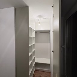 Meble na wymiar Bartosz Dopierała - Biała szafa wnękowa z półkami i drążkiem na ubrania, widok z wnętrza, drewniana podłoga, minimalistyczny styl, oświetlenie żarówką.