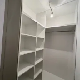 Wnętrze szafy wnękowej z p&oacute;łkami po lewej i drążkiem na ubrania po prawej stronie. Białe wykończenie, oświetlenie punktowe u g&oacute;ry.