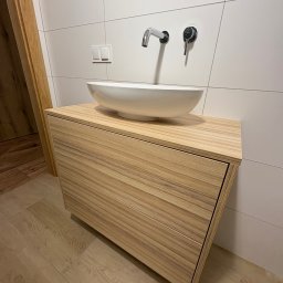 Meble na wymiar Bartosz Dopierała - Nowoczesna łazienka: drewniana szafka pod umywalkę nablatową z chromowaną armaturą na tle białych płytek. Minimalistyczny design, ciepłe oświetlenie.